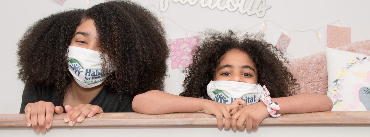 Two girls with dark curly hair look from behind a bunk bed frame wearing Habitat for Humanity masks