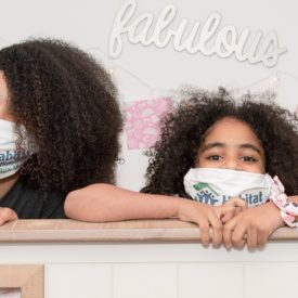 Two girls with dark curly hair look from behind a bunk bed frame wearing Habitat for Humanity masks
