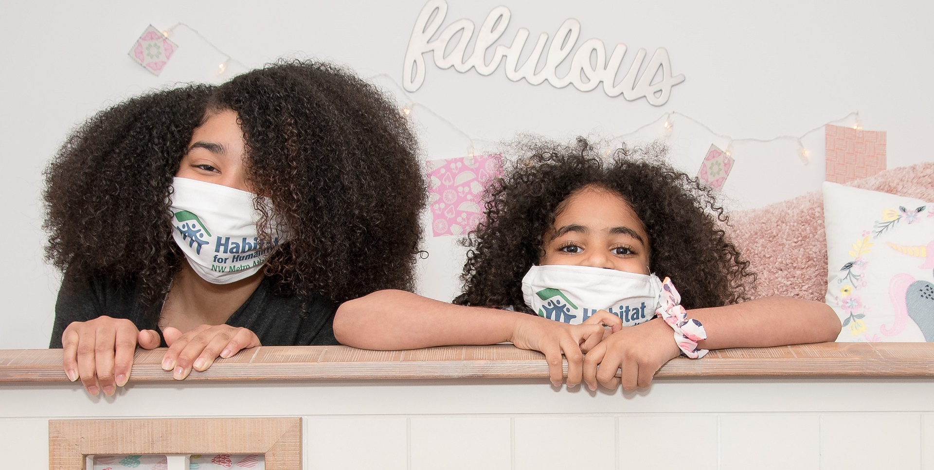 Two girls with dark curly hair look from behind a bunk bed frame wearing Habitat for Humanity masks